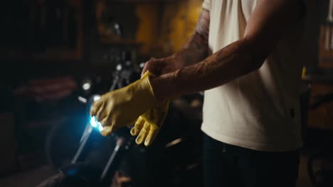 Close-up-of-a-guy-in-a-white-t-shirt-putting-on-yellow-gloves-while-starting-his-working-day-in-a-motorcycle-workshop