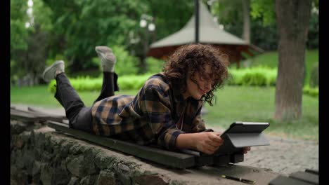 Happy-girl-student-with-curly-hair-in-a-checkered-shirt-uses-a-tablet-and-lies-on-a-bench-in-the-park