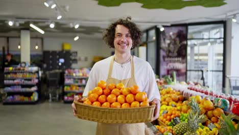 Portrait-of-a-happy-guy-with-curly-hair-as-a-supermarket-worker-who-holds-in-his-hands-a-large-basket-with-a-lot-of-tangerines-and-citrus-fruits-in-the-supermarket