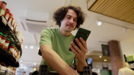 Confident-guy-with-curly-brunette-hair-wearing-a-green-T-shirt-scrolling-through-social-networks-while-shopping-in-a-supermarket-with-a-cart