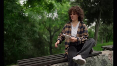 Confident-brunette-girl-with-curly-hair-lies-down-on-a-bench-while-writing-down-her-ideas-and-preparing-for-exams-in-the-park