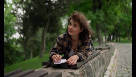 A-thoughtful-brunette-girl-with-curly-hair-writes-down-her-ideas-in-a-notebook-while-lying-on-a-bench-in-the-park