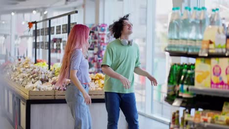 A-happy-girl-with-pink-hair-in-jeans-and-a-brunette-guy-in-a-green-T-shirt-are-dancing-near-the-counter-in-a-supermarket-during-their-shopping