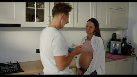 Happy-man-in-white-t-shirt-feeding-his-pregnant-woman-with-fruit-salad-in-the-morning-in-the-kitchen