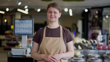 Portrait-of-a-happy-girl-with-blue-eyes-in-a-brown-T-shirt-and-apron-who-stands-near-the-shelves-in-a-supermarket-and-poses-during-her-working-day