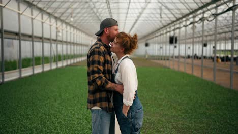 A-loving-couple-a-girl-with-red-hair-and-her-boyfriend-in-a-farmers-cap-stand-among-young-plants-and-sprouts-in-a-greenhouse-on-a-farm