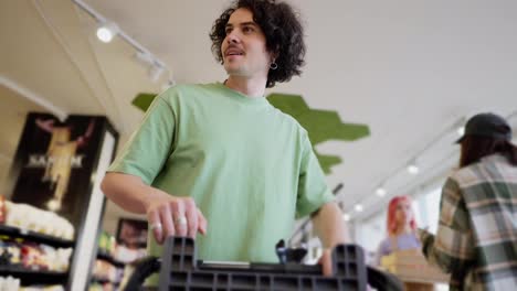 Bottom-view-of-a-confident-brunette-guy-with-curly-hair-in-a-green-T-shirt-carries-a-cart-and-puts-the-goods-he-needs-into-it-in-a-supermarket