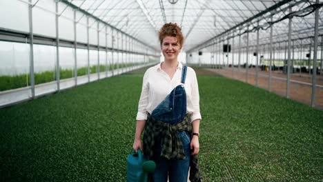 Portrait-of-a-happy-woman-with-red-curly-hair-in-a-white-shirt-who-works-on-a-farm-among-plants-with-a-watering-can-in-her-hands-and-poses