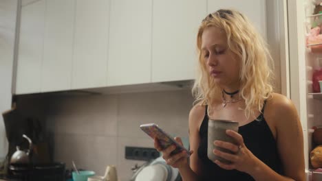 A-tired-blonde-girl-in-a-black-top-drinks-tea-in-the-morning-and-looks-at-her-phone-in-the-morning-in-the-kitchen-near-the-refrigerator