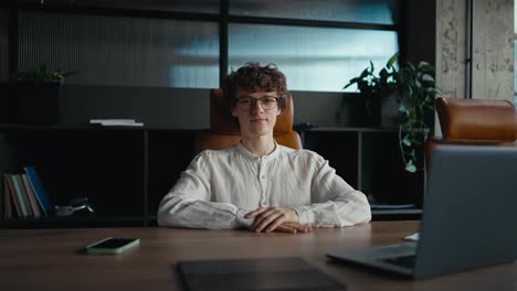 Portrait-of-a-happy-young-guy-with-curly-hair-wearing-glasses-in-a-white-shirt-who-is-sitting-at-a-table-in-the-office-and-posing