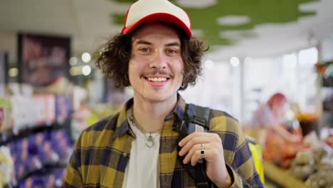 Portrait-of-a-Happy-food-delivery-guy-with-curly-hair-in-a-plaid-shirt-who-stands-in-the-middle-of-a-supermarket-while-choosing-products-for-delivery