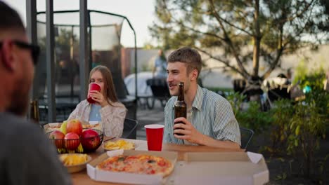 über-Der-Schulter-Hält-Ein-Glücklicher-Blonder-Typ-Eine-Flasche-Bier-In-Den-Händen-Und-Kommuniziert-Aktiv-Mit-Seinen-Freunden,-Während-Er-Beim-Mittagessen-Im-Innenhof-Eines-Landhauses-Am-Tisch-Sitzt