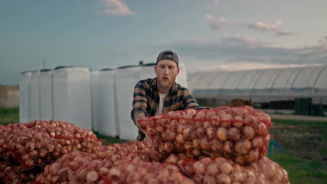 Confident-guy-Farmer-in-a-plaid-shirt-with-a-beard-and-a-cap-shifts-bags-of-onions-while-working-on-the-farm