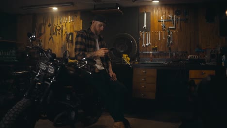 A-mechanic-guy-sits-on-his-moped-and-watches-social-networks-on-his-phone-during-a-break-at-work-in-the-workshop