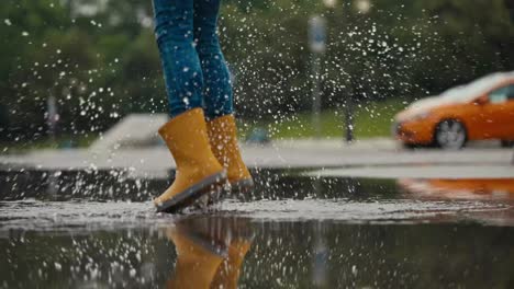 Teenage-girl-in-orange-rubber-boots-energetically-jumps-on-a-puddle-and-splashes-water-while-walking-in-the-park-after-the-rain