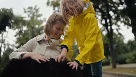 Close-up-of-a-happy-blonde-woman-in-a-white-jacket-petting-her-big-black-dog-with-her-daughter-while-walking-in-the-park-after-the-rain