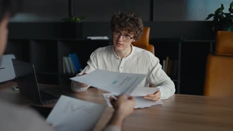 Over-his-shoulder-a-confident-office-employee-shows-reports-and-documents-of-the-current-month-to-a-young-guy-with-curly-hair-while-working-at-his-desk-in-the-office