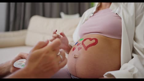 Close-up-a-happy-man-uses-a-palette-to-paint-on-the-belly-of-his-pregnant-wife-during-their-leisure-time-and-waiting-for-the-baby-at-home