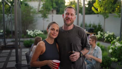 Portrait-of-a-happy-blond-guy-with-a-beard-in-a-gray-T-shirt-who-hugs-his-girlfriend-and-poses-while-relaxing-with-company-in-the-courtyard-of-a-country-house