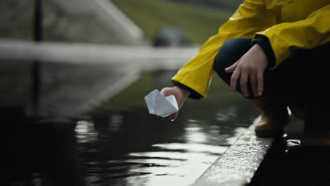 Close-up-a-teenage-girl-in-a-yellow-jacket-launches-a-white-paper-boat-into-the-water-through-a-large-puddle-during-the-rain-on-the-street