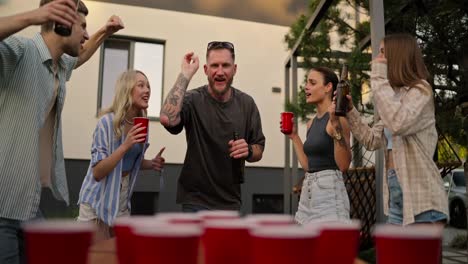 Close-up-of-a-happy-blond-guy-with-a-bottle-of-beer-in-his-hands-hitting-a-small-table-tennis-ball-into-a-red-cup-during-a-party-in-the-courtyard-of-a-country-house