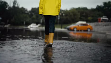 Vista-Trasera-De-Una-Adolescente-Con-Una-Chaqueta-Amarilla-Y-Botas-De-Goma-Naranjas-Caminando-Por-Un-Gran-Charco-Y-Empujando-Agua-Mientras-Camina-Después-De-La-Lluvia-En-La-Calle.