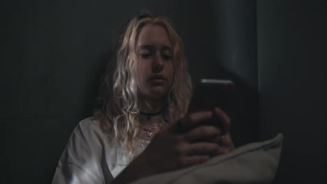 A-blonde-girl-in-a-white-T-shirt-leans-against-a-gray-wall-and-watches-social-networks-before-going-to-bed-late-at-night