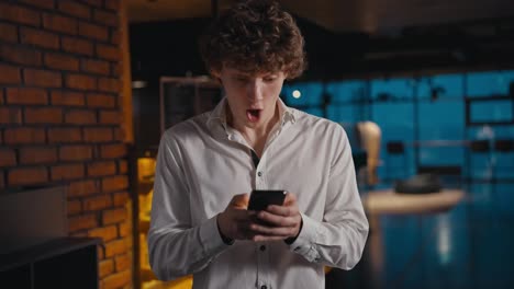 A-young-guy-with-curly-hair-is-pleasantly-surprised-and-jumps-with-happiness-when-he-looks-at-the-screen-of-his-phone-in-the-office