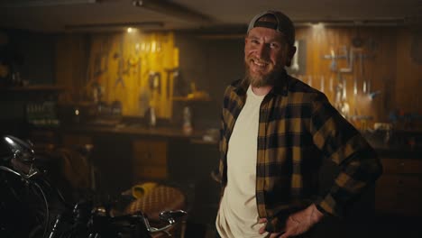 Portrait-of-a-cheerful-guy-mechanic-in-a-checkered-shirt-and-cap-posing-and-laughing-in-his-workshop-studio