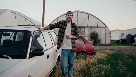 Portrait-of-a-happy-farmer-guy-in-a-cap-and-a-plaid-shirt-posing-near-a-car-among-greenhouses-and-fields-on-a-farm