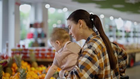 Primer-Plano-De-Una-Chica-Morena-Segura-De-Sí-Misma-Con-Una-Camisa-A-Cuadros-Y-Su-Pequeño-Hijo-En-Brazos-Examinando-Frutas-En-El-Departamento-Del-Supermercado.