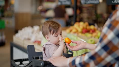 Nahaufnahme,-Ein-Kleines-Mädchen-Bekommt-Von-Ihrer-Mutter-Eine-Mandarine-Und-Schaut-Sie-Sich-Beim-Einkaufen-In-Einem-Supermarkt-An