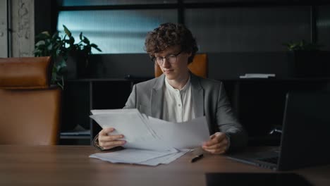 From-the-side-a-confident-young-guy-with-curly-hair-and-glasses-in-a-gray-jacket-examines-documents-and-papers-while-sitting-at-a-table-in-a-modern-office