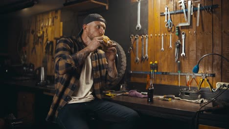 Un-Mecánico-Feliz-Con-Camisa-A-Cuadros-Come-Una-Hamburguesa-Y-Bebe-Refresco-Durante-El-Almuerzo-Y-El-Descanso-En-Su-Taller-De-Trabajo