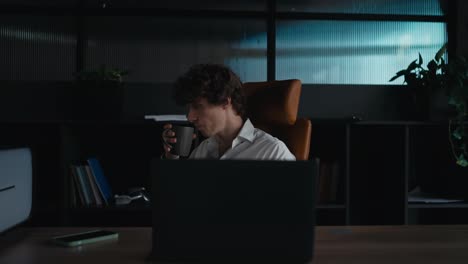 A-young-guy-with-curly-hair-in-a-white-shirt-sits-at-a-table-and-works-on-a-laptop-while-drinking-coffee-in-the-office