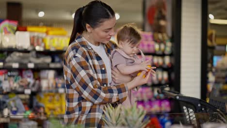 Una-Chica-Morena-Segura-De-Sí-Misma-Con-Una-Camisa-A-Cuadros-Le-Muestra-Mandarinas-A-Su-Pequeña-Hija-Y-Las-Examina-Mientras-Elige-Frutas-En-Un-Supermercado-Mientras-Hace-Compras.