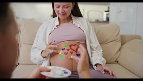 A-happy-pregnant-brunette-woman-draws-hearts-on-her-belly-with-her-husband-using-bright-colors-while-expecting-her-baby