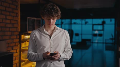 Confident-guy-with-curly-hair-in-a-white-shirt-typing-on-his-phone-and-standing-in-the-evening-office