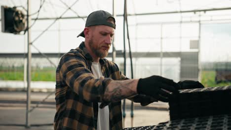 Confident-guy-Farmer-with-a-beard-in-a-cap-in-a-plaid-shirt-arranges-boxes-for-seedlings-and-inspects-them-during-his-work-in-a-greenhouse-on-a-farm