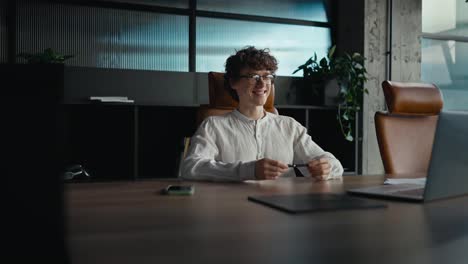 Vista-Lateral-De-Un-Joven-Feliz-Con-Cabello-Rizado-Que-Usa-Anteojos-Y-Una-Camisa-Blanca-Se-Comunica-Por-Videoconferencia-Usando-Una-Computadora-Portátil-Gris-Mientras-Está-Sentado-En-Una-Mesa-En-La-Oficina.