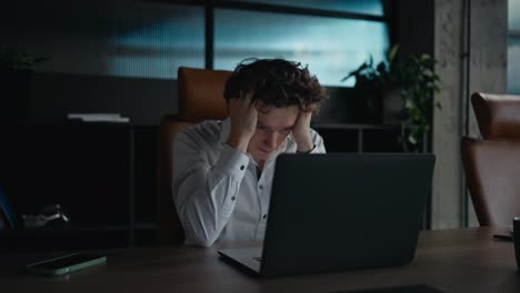 A-tired-guy-with-curly-hair-in-a-white-shirt-looks-tiredly-at-a-gray-laptop-and-holds-his-head-with-his-hands-during-a-hard-day-at-work-in-the-office