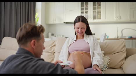 Over-his-shoulder-a-man-in-a-gray-shirt-paints-with-bright-colors-on-the-belly-of-his-pregnant-wife-at-home-in-a-modern-apartment