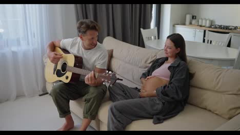 Glücklicher-Mann-Mittleren-Alters-Im-Weißen-T-Shirt,-Der-Gitarre-Spielt,-Während-Er-Am-Wochenende-Mit-Seiner-Schwangeren-Brünetten-Frau-Zu-Hause-Auf-Dem-Sofa-Abhängt