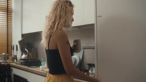 A-young-blonde-girl-in-a-black-top-with-a-tattoo-drinks-tea-in-the-morning-and-looks-into-the-refrigerator-with-a-phone-in-her-hands-in-the-morning