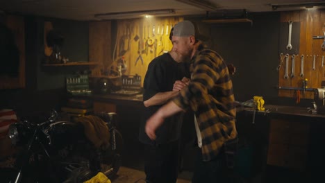 Happy-brunette-guy-and-a-mechanic-in-a-checkered-shirt-shake-hands-after-successfully-fixing-a-motorcycle-in-a-workshop