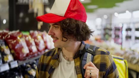 Un-Repartidor-De-Comida-Confiado,-Con-Una-Gorra-Roja-Y-Blanca,-Camina-Cerca-De-Las-Filas-De-Comestibles-Y-Coloca-Los-Productos-Que-Necesita-En-Una-Bolsa-Amarilla-Antes-De-La-Entrega.