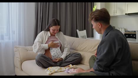 Happy-brunette-pregnant-woman-tries-on-clothes-for-her-baby-whose-child-they-are-expecting-together-with-her-husband-at-home-on-the-sofa