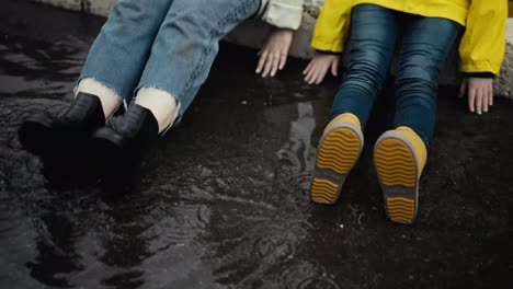 Close-up-shot-of-a-woman-in-black-rubber-boots-and-her-teenage-daughter-in-orange-rubber-boots-putting-their-feet-on-a-puddle-after-rain-on-the-street