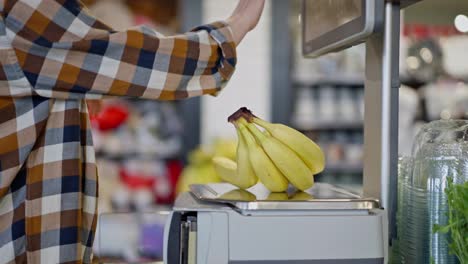 Close-up-of-a-confident-woman-in-a-plaid-shirt-weighing-bananas-on-an-electronic-scale-in-a-supermarket-while-shopping-with-her-infant-daughter