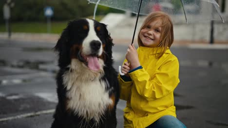 Portrait-of-a-happy-blonde-teenage-girl-in-a-yellow-jacket-and-with-an-umbrella-who-poses-together-with-her-black-and-white-rain-dog-in-the-park-during